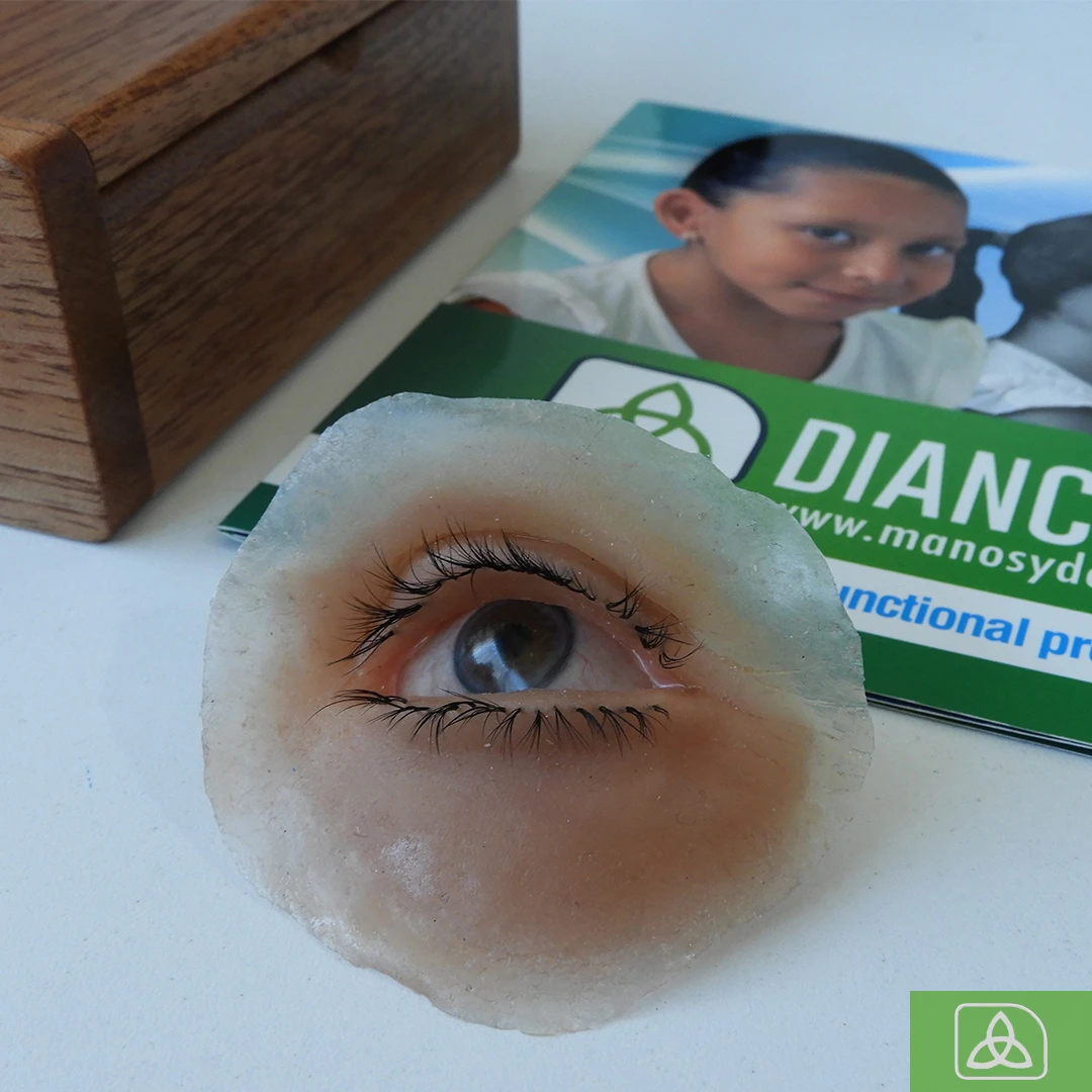 Eye prosthesis resting on white counter. In the background is a wooden case and an informative brochure with the Dianceht logo.